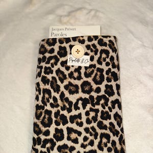 Peut inclure: Une housse rectangulaire avec un motif léopard beige et marron. La housse est fermée par un bouton en bois. Le texte "Jacques Prévert Paroles" est visible sur une carte blanche. La marque "Piplette & Co" est également visible.