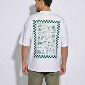 Peut inclure: Un t-shirt blanc à manches courtes avec un grand motif rectangulaire dans le dos. Le motif est encadré par une bordure à damier vert et blanc et contient diverses petites illustrations au trait vert, incluant des voitures, des animaux et du texte.