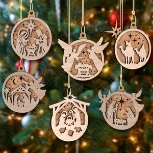 May include: Six wooden Christmas ornaments with religious themes. Each round ornament features intricate cut-out designs depicting nativity scenes, angels, and stars. They hang from gold strings against a blurred Christmas tree background.