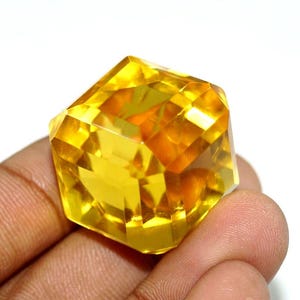May include: A faceted, golden-yellow gemstone with a geometric shape. The gem is held in a hand, showcasing its brilliant cut and clarity. The gemstone's facets reflect light, creating a dazzling display of colour and brilliance.
