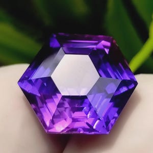 May include: A faceted, hexagonal gemstone with a central white facet and gradient purple hues. The gem's facets reflect light, creating a sparkling effect. The gemstone is held against a blurred green and white background.