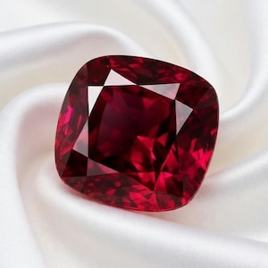 Puede incluir: Un gran rubí tallado en cojín. La gema roja oscura se encuentra sobre una tela de satén blanco. Las facetas de la gema reflejan la luz, creando un brillo brillante. Una pieza de joyería de lujo.