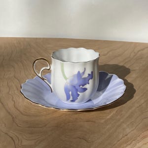 Puede incluir: Juego de taza y platillo blancos con diseño floral azul. La taza tiene asa y borde dorados, y el platillo tiene borde festoneado. El diseño floral es un iris. El conjunto está sobre una superficie de madera.