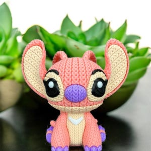 May include: A knitted figurine of a cartoon character in shades of pink, purple, and beige. The character has large ears, black eyes, and a purple nose. The figurine is set against a blurred background of green plants.