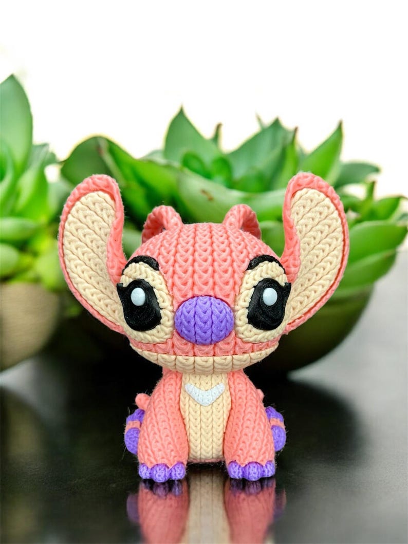 May include: A knitted figurine of a cartoon character in shades of pink, purple, and beige. The character has large ears, black eyes, and a purple nose. The figurine is set against a blurred background of green plants.