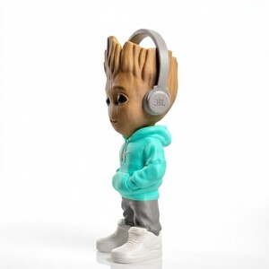 Op de afbeelding: Een beeldje van een personage met een houten uiterlijk, gekleed in een turquoise hoodie, grijze broek en witte sneakers. Het figuur heeft een grijze koptelefoon met het JBL-logo. De achtergrond is wit.
