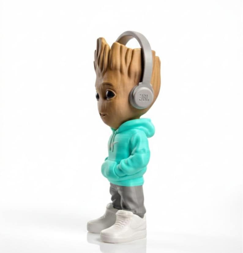 Op de afbeelding: Een beeldje van een personage met een houten uiterlijk, gekleed in een turquoise hoodie, grijze broek en witte sneakers. Het figuur heeft een grijze koptelefoon met het JBL-logo. De achtergrond is wit.