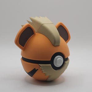 May include: A spherical orange figurine featuring a tan mohawk-style crest, two dark brown ears, and a central circular button. The design includes a black horizontal band and a tan jagged pattern near the base, resembling a stylised creature character.