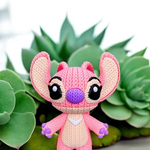 May include: A pink and purple figurine of the character Angel from the Disney movie Lilo & Stitch. The figurine has large black eyes, a purple nose, and pink ears. The figure is standing in front of green plants.