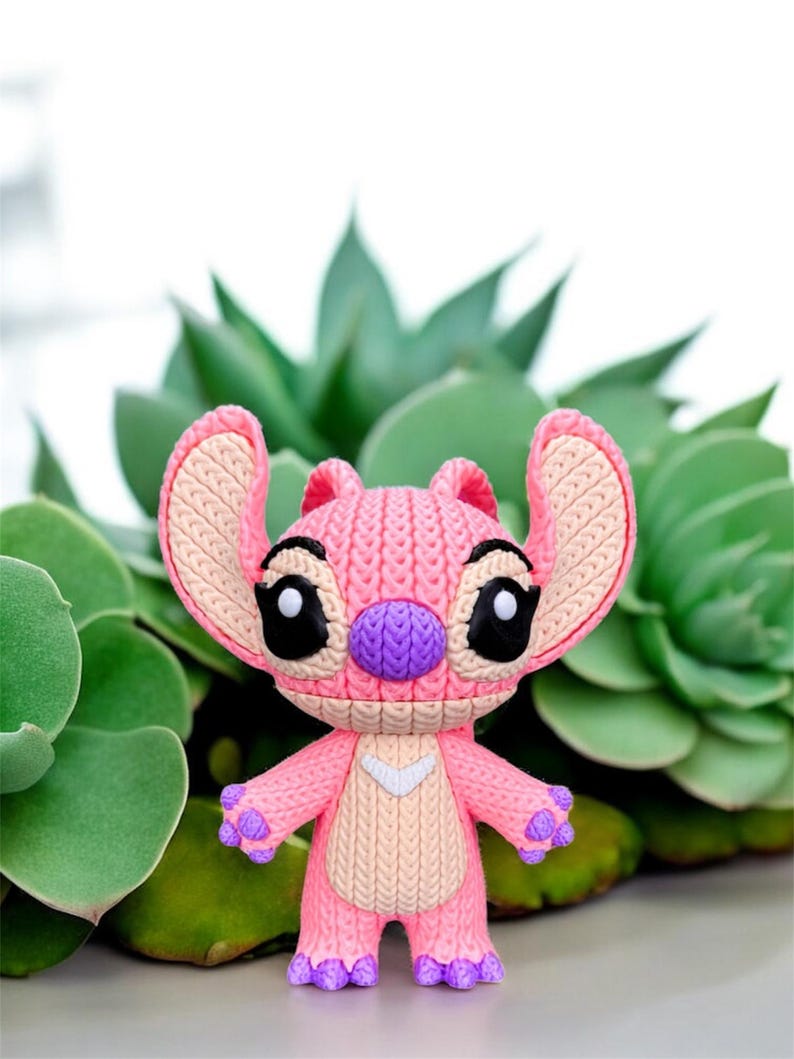 May include: A pink and purple figurine of the character Angel from the Disney movie Lilo & Stitch. The figurine has large black eyes, a purple nose, and pink ears. The figure is standing in front of green plants.