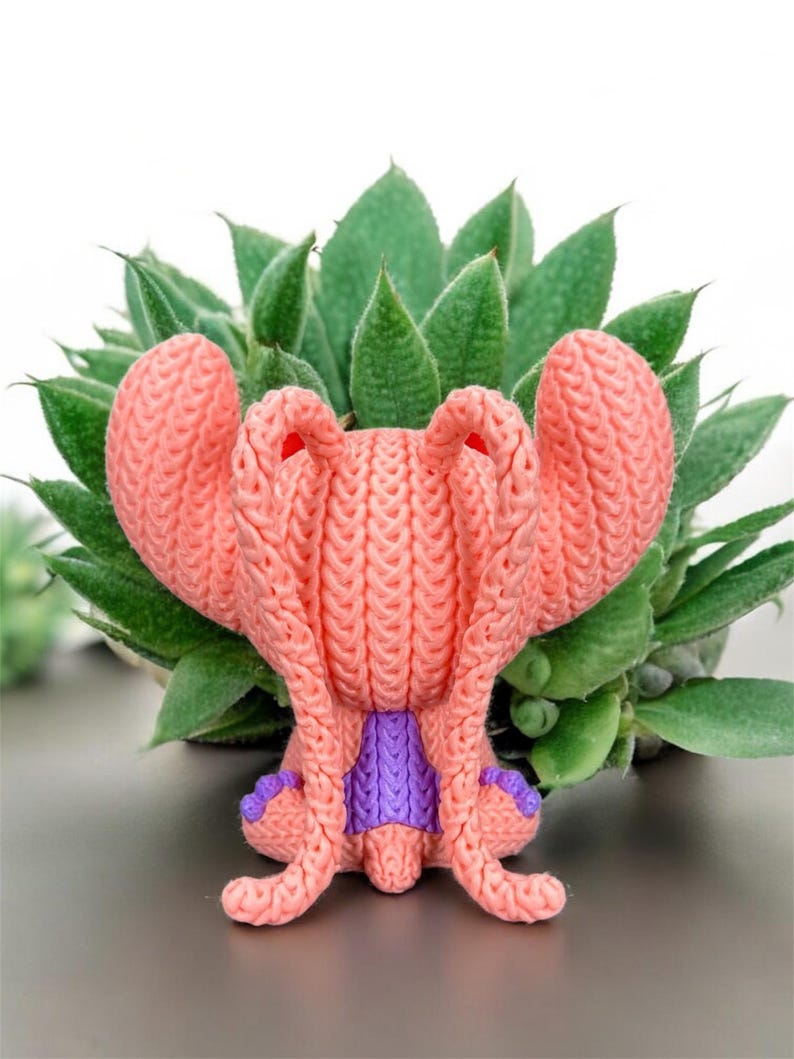 May include: A handmade, coral-colored knitted creature with large, textured ears and a purple accent. The whimsical figure is set against a backdrop of green succulent leaves, creating a playful contrast of textures and colors.