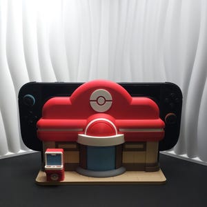 May include: A red and white 3D-printed model of a building with a Poké Ball logo, designed to hold a Nintendo Switch. The model has a small, matching vending machine. The background is a black surface and a white wavy wall.