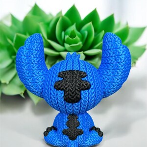 May include: A blue figurine of a cartoon character with large ears and black accents. The figurine has a textured appearance, with the black accents on the chest and head. The background features a green succulent plant.