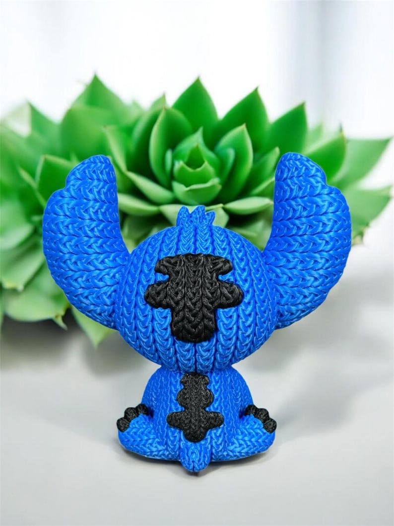 May include: A blue figurine of a cartoon character with large ears and black accents. The figurine has a textured appearance, with the black accents on the chest and head. The background features a green succulent plant.