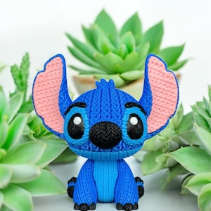 May include: A blue Stitch figurine with large ears and black eyes. The figurine has a knitted texture and is surrounded by green succulents. The ears are pink on the inside.