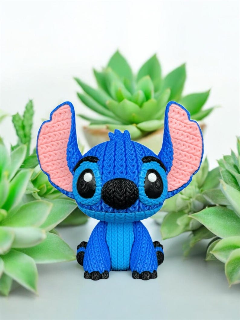 May include: A blue Stitch figurine with large ears and black eyes. The figurine has a knitted texture and is surrounded by green succulents. The ears are pink on the inside.