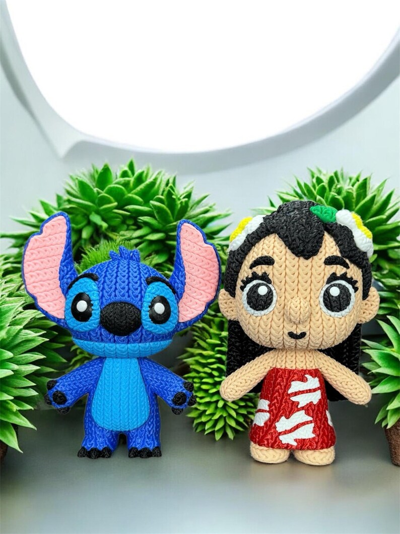 May include: Two knitted figurines of characters from the animated film Lilo & Stitch. One is blue with large ears, the other is a girl in a red dress. They are set against a backdrop of green plants.