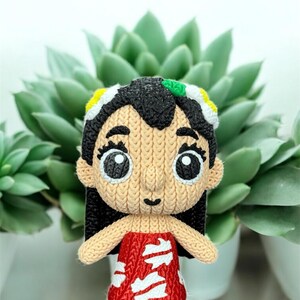 May include: A handmade, knitted doll with black hair, large eyes, and a red dress with white accents. The doll has a tan face and arms, and is wearing a headband with yellow, white, and green details. The doll is standing in front of a green succulent plant.