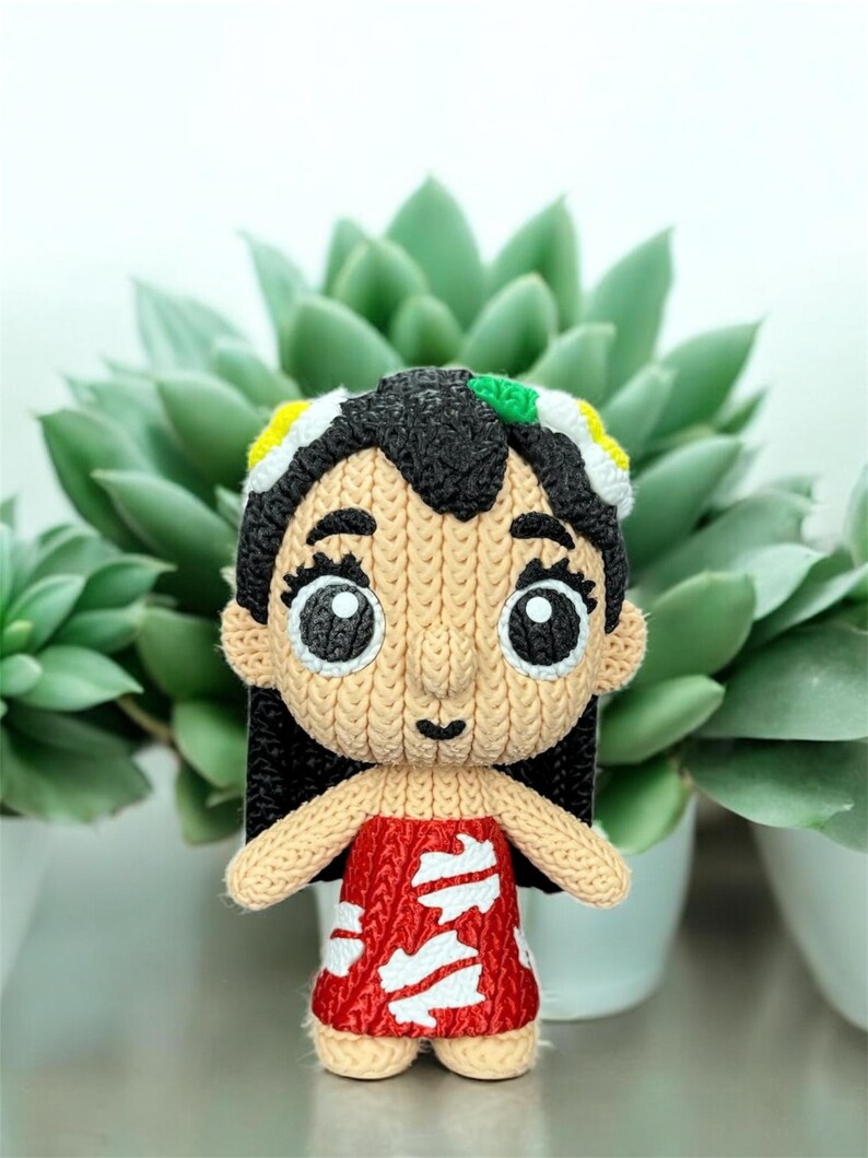 May include: A handmade, knitted doll with black hair, large eyes, and a red dress with white accents. The doll has a tan face and arms, and is wearing a headband with yellow, white, and green details. The doll is standing in front of a green succulent plant.