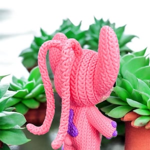 May include: A pink, knitted figurine of a cartoon character with long, looped ears and purple accents. The figure is set against a backdrop of green succulents in terracotta pots, creating a playful contrast of textures and colors.