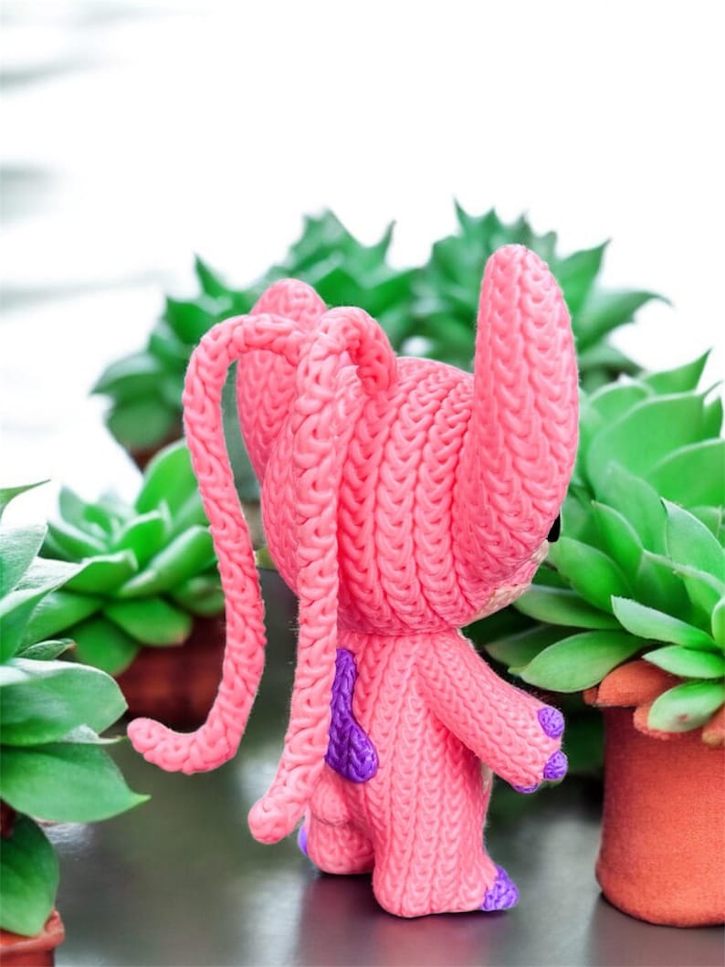 May include: A pink, knitted figurine of a cartoon character with long, looped ears and purple accents. The figure is set against a backdrop of green succulents in terracotta pots, creating a playful contrast of textures and colors.