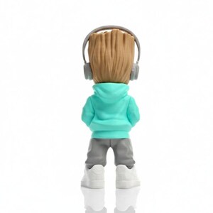 Op de afbeelding: Een beeldje van een personage met een houten hoofd, gekleed in een turquoise hoodie, grijze broek, witte sneakers en een grijze koptelefoon. De figuur staat met de rug naar de kijker toe, tegen een witte achtergrond. Het hoofd van de figuur is bruin en getextureerd.