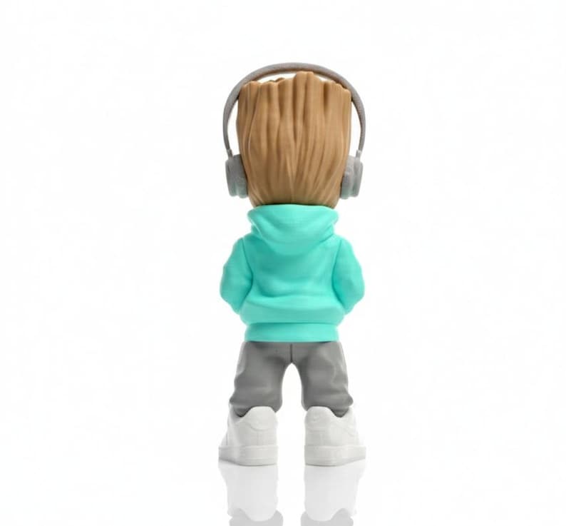 Op de afbeelding: Een beeldje van een personage met een houten hoofd, gekleed in een turquoise hoodie, grijze broek, witte sneakers en een grijze koptelefoon. De figuur staat met de rug naar de kijker toe, tegen een witte achtergrond. Het hoofd van de figuur is bruin en getextureerd.