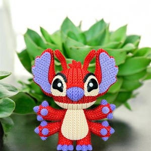 May include: A red and purple Stitch character figurine with a cream-colored belly. The figurine has large black eyes and is set against a backdrop of green foliage. The character's ears and feet are purple.