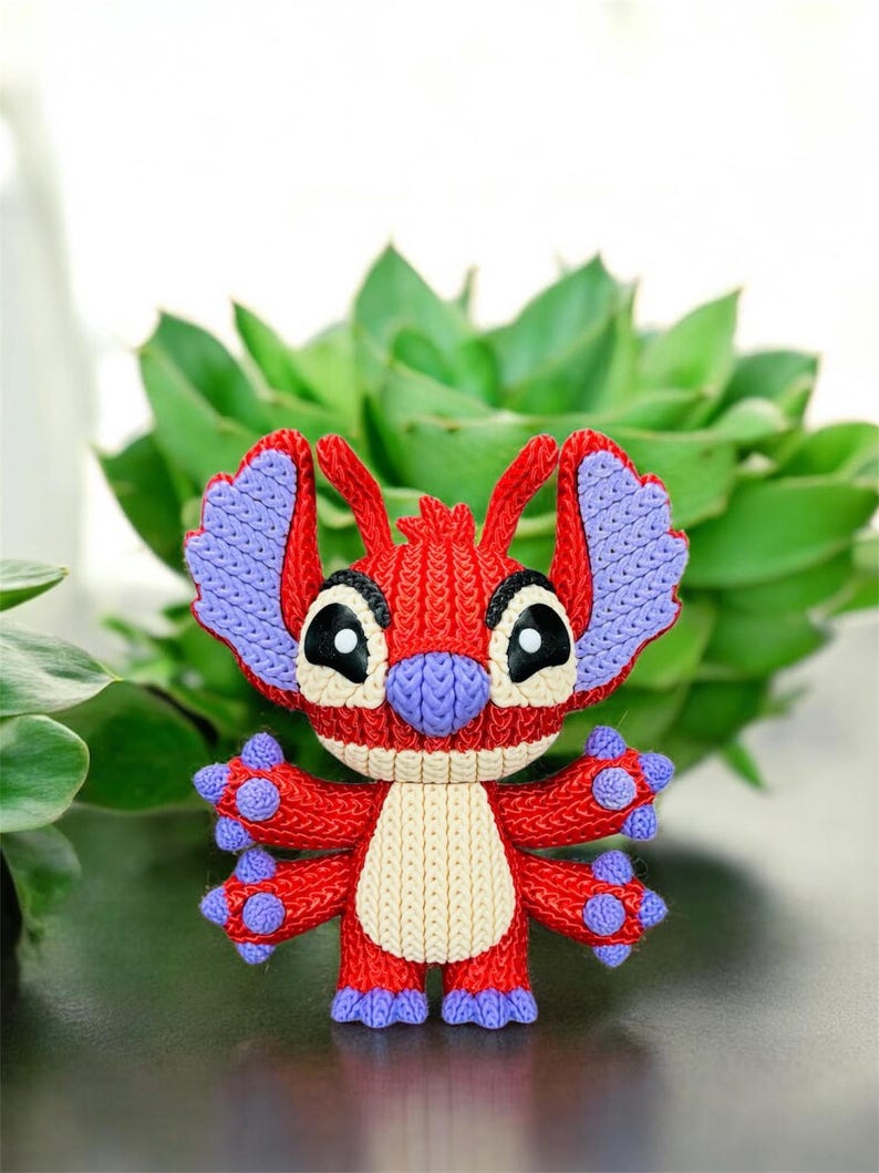 May include: A red and purple Stitch character figurine with a cream-colored belly. The figurine has large black eyes and is set against a backdrop of green foliage. The character's ears and feet are purple.