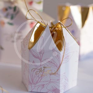 K&ouml;nnte beinhalten: Eine wei&szlig;e, sechseckige Geschenkbox mit goldenem Deckel und rosa Blumenmuster. Die Box hat eine goldene Schnur und den goldenen Schriftzug "Happy Mother's Day".