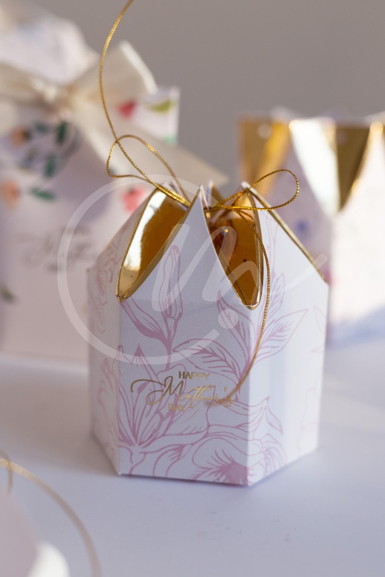 K&ouml;nnte beinhalten: Eine wei&szlig;e, sechseckige Geschenkbox mit goldenem Deckel und rosa Blumenmuster. Die Box hat eine goldene Schnur und den goldenen Schriftzug "Happy Mother's Day".