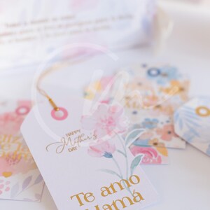 K&ouml;nnte beinhalten: Wei&szlig;e Geschenkanh&auml;nger mit floralen Designs und goldfarbenen Buchstaben. Ein Anh&auml;nger tr&auml;gt die Aufschrift "Happy Mother's Day" und "Te amo Mam&aacute;" mit einer rosa Blumenillustration. Andere Anh&auml;nger zeigen Aquarell-Blumenmuster.