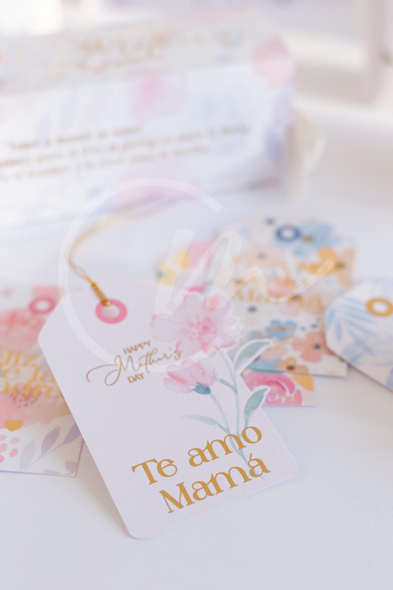 K&ouml;nnte beinhalten: Wei&szlig;e Geschenkanh&auml;nger mit floralen Designs und goldfarbenen Buchstaben. Ein Anh&auml;nger tr&auml;gt die Aufschrift "Happy Mother's Day" und "Te amo Mam&aacute;" mit einer rosa Blumenillustration. Andere Anh&auml;nger zeigen Aquarell-Blumenmuster.