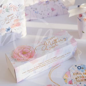 K&ouml;nnte beinhalten: Geschenkboxen und Anh&auml;nger mit floralen Designs und dem Text "Happy Mother's Day". Die Boxen und Anh&auml;nger sind mit rosa und blauen Blumen und goldfarbenen Buchstaben verziert. Ein rosa Blumenaufkleber befindet sich auf der Box.