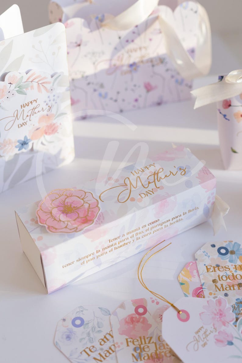 K&ouml;nnte beinhalten: Geschenkboxen und Anh&auml;nger mit floralen Designs und dem Text "Happy Mother's Day". Die Boxen und Anh&auml;nger sind mit rosa und blauen Blumen und goldfarbenen Buchstaben verziert. Ein rosa Blumenaufkleber befindet sich auf der Box.