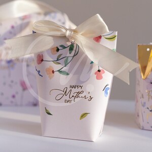 K&ouml;nnte beinhalten: Eine kleine, hellrosa Geschenkbox mit einer cremefarbenen Schleife. Die Box ist mit einem Blumenmuster und den Worten "Happy Mother's Day" in goldener Schrift verziert. Weitere Boxen sind im Hintergrund zu sehen.