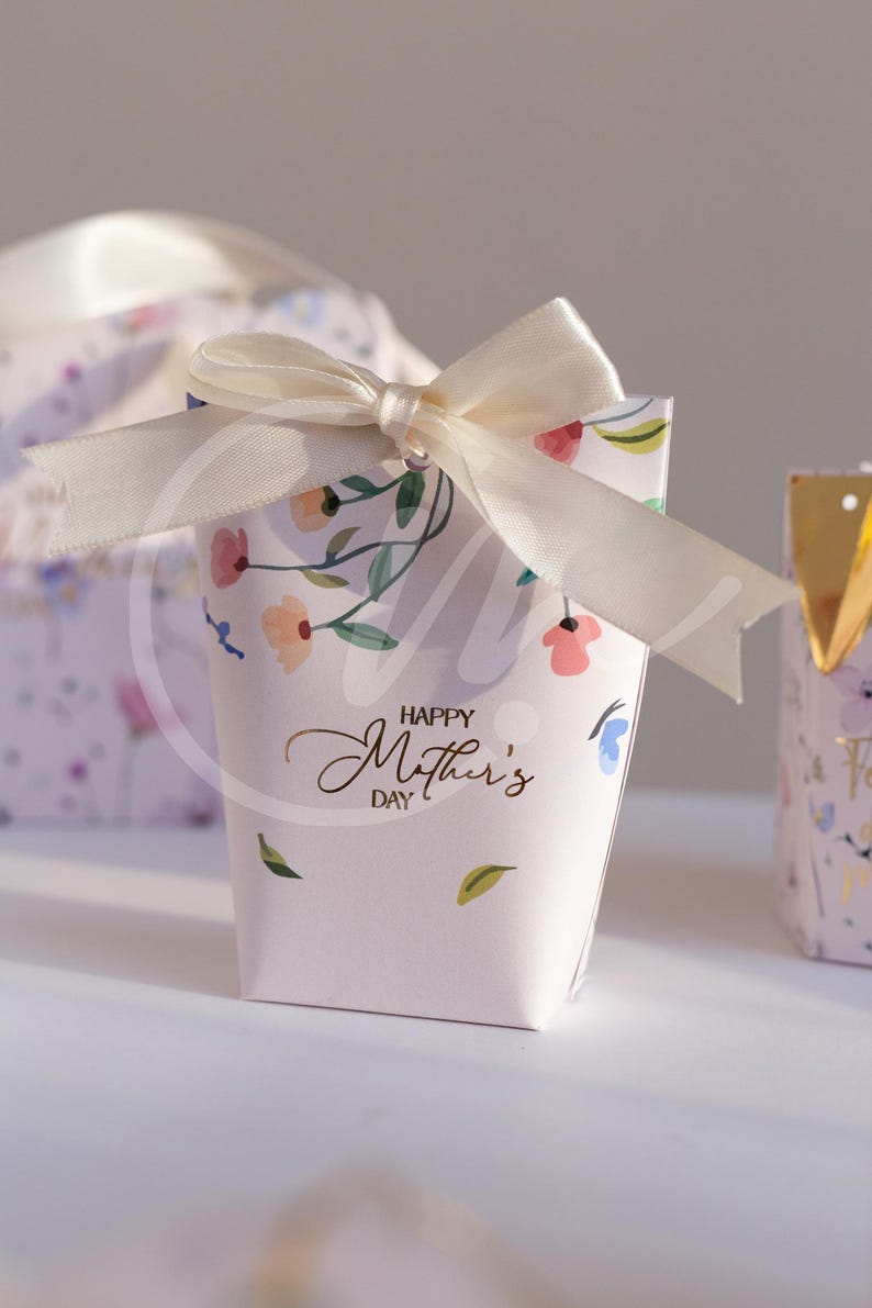 K&ouml;nnte beinhalten: Eine kleine, hellrosa Geschenkbox mit einer cremefarbenen Schleife. Die Box ist mit einem Blumenmuster und den Worten "Happy Mother's Day" in goldener Schrift verziert. Weitere Boxen sind im Hintergrund zu sehen.