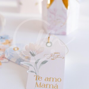 K&ouml;nnte beinhalten: Ein Geschenkanh&auml;nger mit einem Aquarell-Blumenmuster und dem Text "Happy Mother's Day" und "Te amo Mam&aacute;" in goldfarbenen Buchstaben. Der Anh&auml;nger ist an einer Geschenkbox mit goldenem Band befestigt.