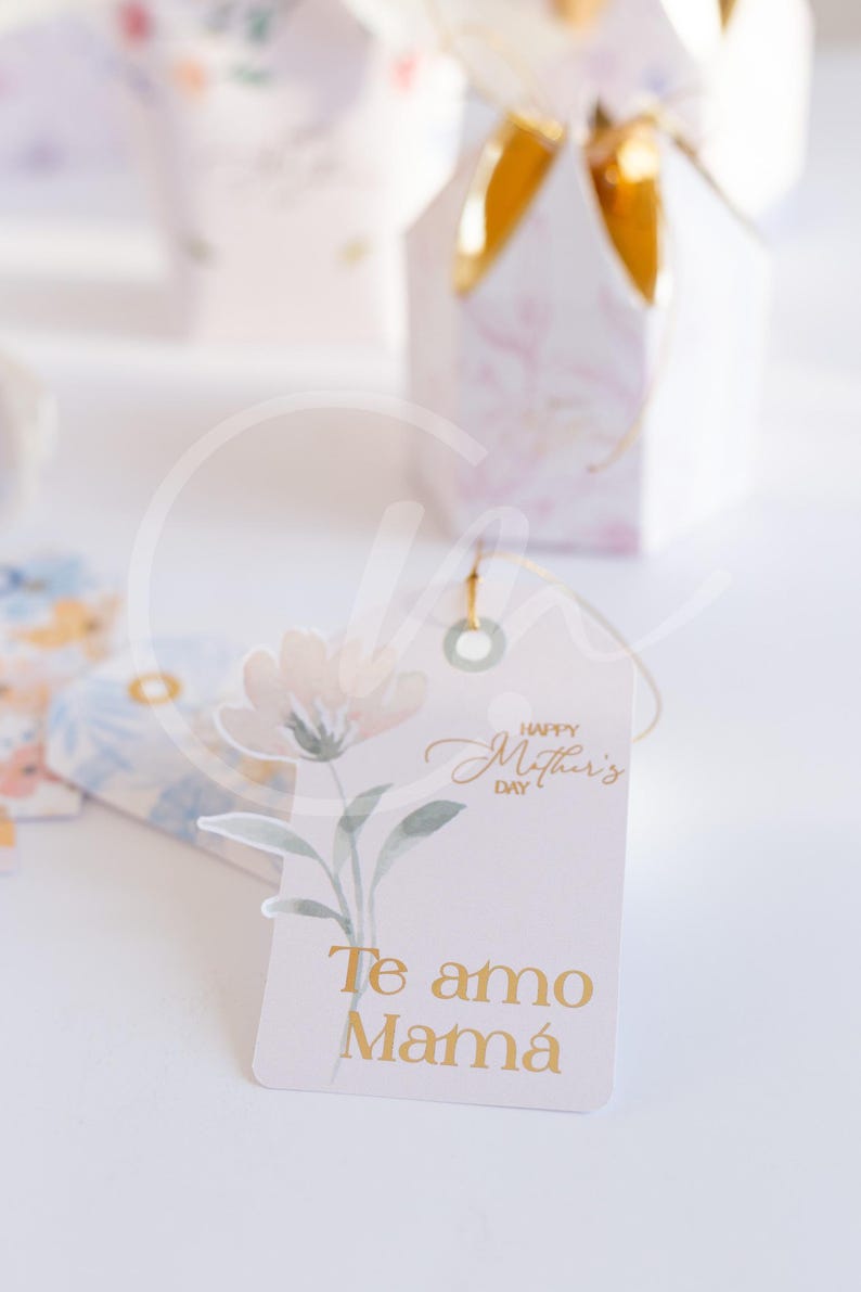 K&ouml;nnte beinhalten: Ein Geschenkanh&auml;nger mit einem Aquarell-Blumenmuster und dem Text "Happy Mother's Day" und "Te amo Mam&aacute;" in goldfarbenen Buchstaben. Der Anh&auml;nger ist an einer Geschenkbox mit goldenem Band befestigt.