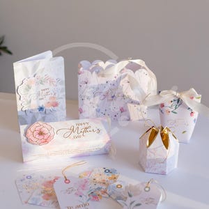 Puede incluir: Surtido de regalos para el Día de la Madre, que incluyen tarjetas, cajas de regalo y etiquetas. Los artículos presentan diseños florales y las frases "Happy Mother's Day" y "Te amo Mama". La paleta de colores incluye pasteles y detalles dorados.