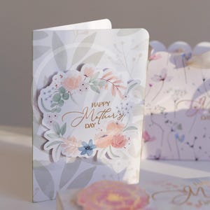 K&ouml;nnte beinhalten: Ein Gru&szlig;karten- und Geschenkbox-Set zum Muttertag. Die Karte zeigt ein florales Kranzdesign mit dem goldenen Schriftzug "Happy Mother's Day". Die Geschenkbox hat ein passendes Blumenmuster.