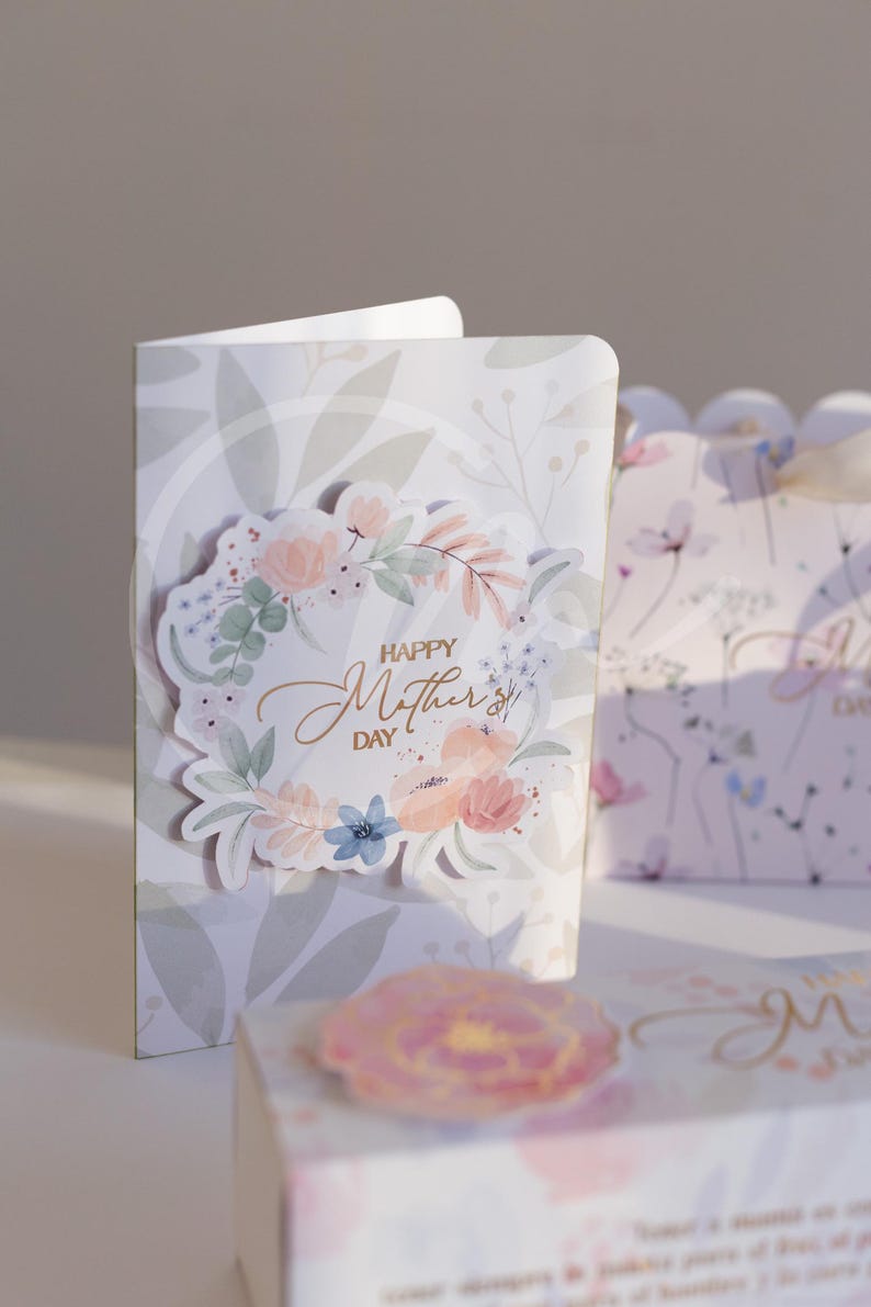 K&ouml;nnte beinhalten: Ein Gru&szlig;karten- und Geschenkbox-Set zum Muttertag. Die Karte zeigt ein florales Kranzdesign mit dem goldenen Schriftzug "Happy Mother's Day". Die Geschenkbox hat ein passendes Blumenmuster.