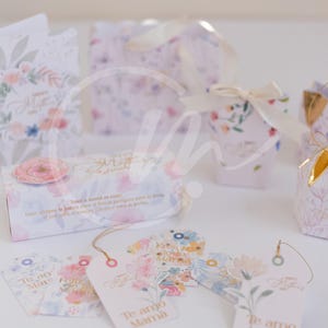 K&ouml;nnte beinhalten: Geschenkboxen und Anh&auml;nger mit floralen Designs. Die Boxen und Anh&auml;nger sind mit pastellfarbenen Blumen und den Texten "Happy Mother's Day" und "Te amo Mam&aacute;" verziert. Die Boxen sind mit B&auml;ndern gebunden.
