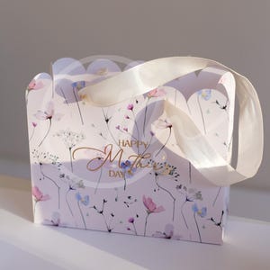K&ouml;nnte beinhalten: Eine Geschenkbox mit floralem Muster in Rosa, Lila und Blau, mit wei&szlig;em Schleifenhenkel. Auf der Vorderseite der Tasche steht in goldener Schrift "Happy Mother's Day".