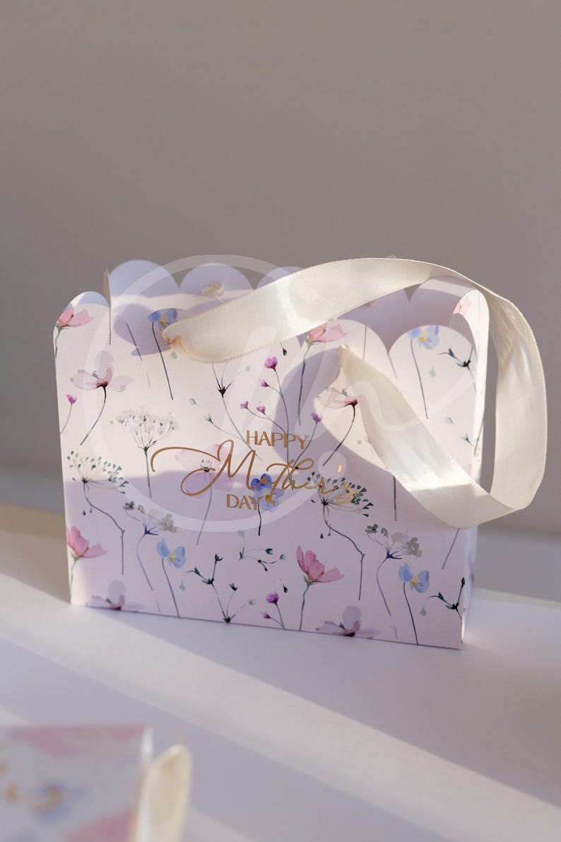 K&ouml;nnte beinhalten: Eine Geschenkbox mit floralem Muster in Rosa, Lila und Blau, mit wei&szlig;em Schleifenhenkel. Auf der Vorderseite der Tasche steht in goldener Schrift "Happy Mother's Day".