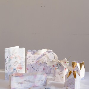 K&ouml;nnte beinhalten: Eine Auswahl an Geschenkboxen, Anh&auml;ngern und Karten mit floralen Designs und dem Text "Happy Mother's Day". Die Boxen sind wei&szlig; mit goldenen Akzenten und Schleifen. Die Karten und Anh&auml;nger haben Blumenmuster und Text in mehreren Sprachen.