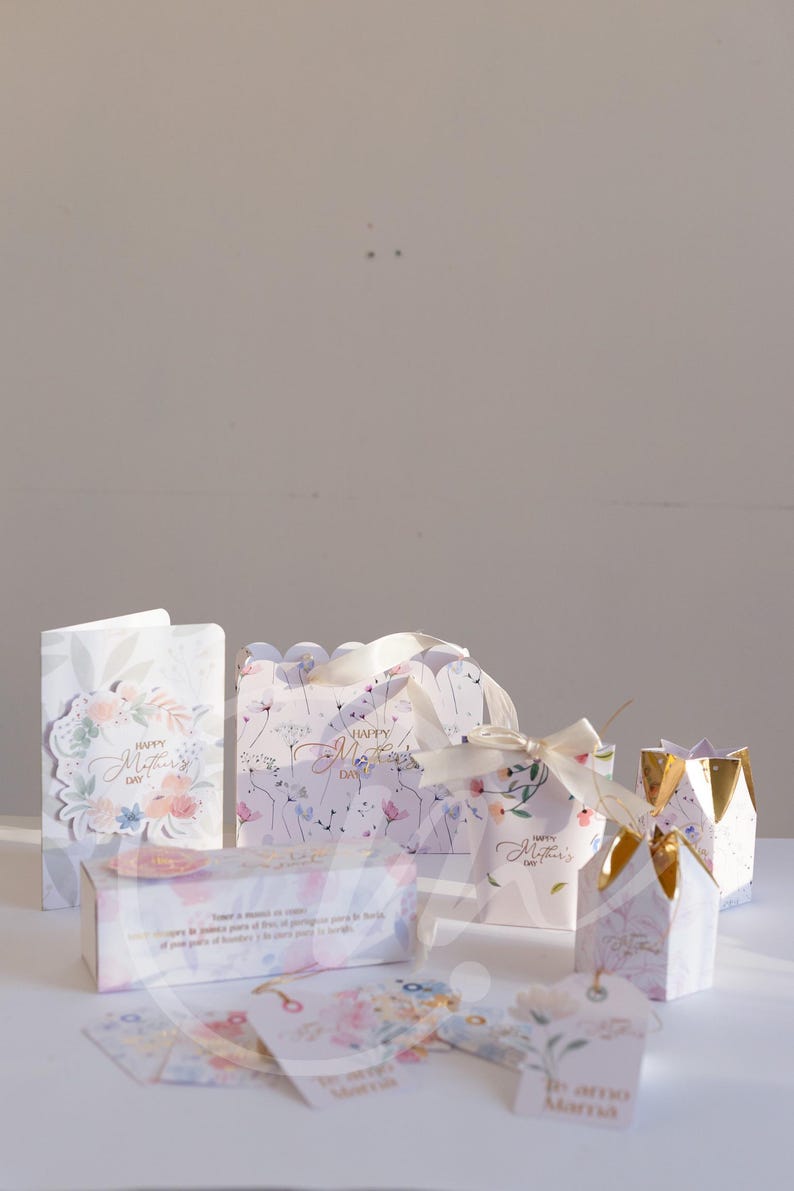 K&ouml;nnte beinhalten: Eine Auswahl an Geschenkboxen, Anh&auml;ngern und Karten mit floralen Designs und dem Text "Happy Mother's Day". Die Boxen sind wei&szlig; mit goldenen Akzenten und Schleifen. Die Karten und Anh&auml;nger haben Blumenmuster und Text in mehreren Sprachen.