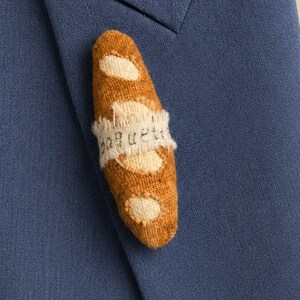 May include: A brown fabric baguette-shaped brooch pinned to the lapel of a blue blazer. The brooch features a cream-coloured fabric band wrapped around the centre with the word baguette printed in black ink on the fabric strip.