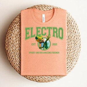 May include: Peach-colored t-shirt with the word "ELECTRO" in green block letters. Below the word is a cartoon character and the text "EST 2021 SPIDEY AND HIS AMAZING FRIENDS". The shirt is folded on a woven, circular surface.