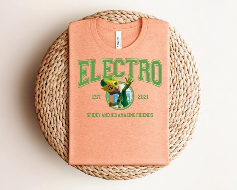 May include: Peach-colored t-shirt with the word "ELECTRO" in green block letters. Below the word is a cartoon character and the text "EST 2021 SPIDEY AND HIS AMAZING FRIENDS". The shirt is folded on a woven, circular surface.